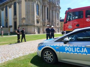 oznakowany radiowóz, a za nim dwóch umundurowanych policjantów ruchu drogowego stojących tyłem. W tle bazylika w Krzeszowie