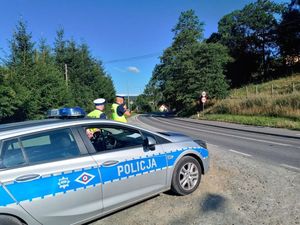 dwóch umundurowanych policjantów ruchu drogowego stoi przed oznakowanym radiowozem, jeden z nich wykonuje pomiar prędkości
