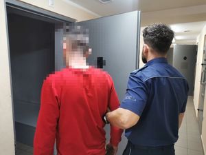 Mężczyzna w czerwonej bluzie, którego za ramię trzyma umundurowany policjant